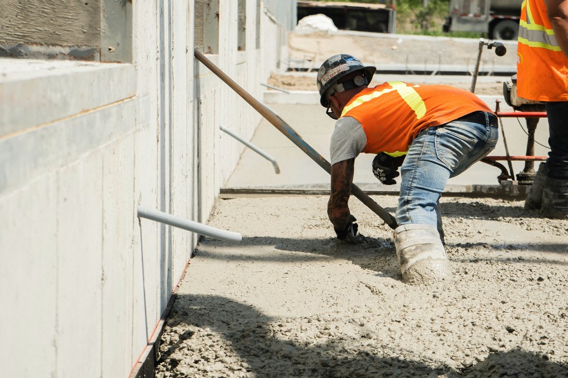 A worker in a high-visibility vest spreading fresh concrete with a rake along a formed slab edge.