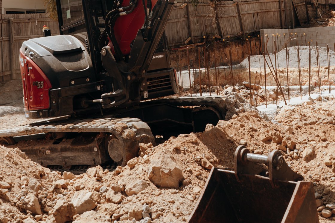 A tracked excavator digging a footing trench, with steel reinforcing bar set in the excavation and the bucket resting in loose soil.