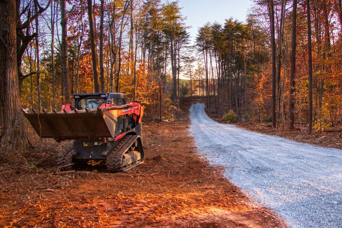 A compact tracked loader with a raised bucket parked beside a freshly graded gravel driveway cut through autumn woods.